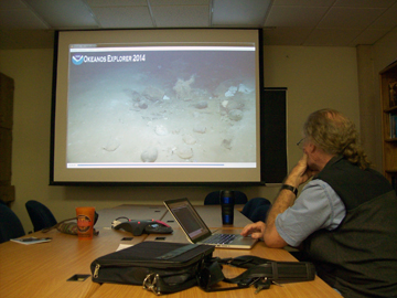 A man seated at a table in front of a large screen TV with a live feed of an underwater expedition.