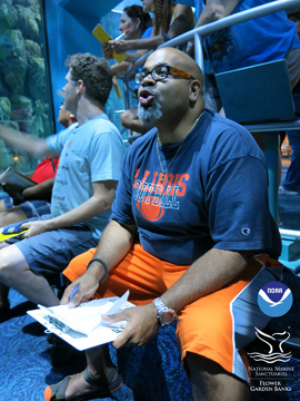 Teachers sitting on a bench with others standing behind them as they look at a large aquarium full of fish.