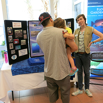 A father holds a young child while talking to a volunteer in front of a table display about conservation