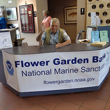A man waves from behind the entry desk with a Flower Garden Banks National Marine Sanctuary banner in front of him