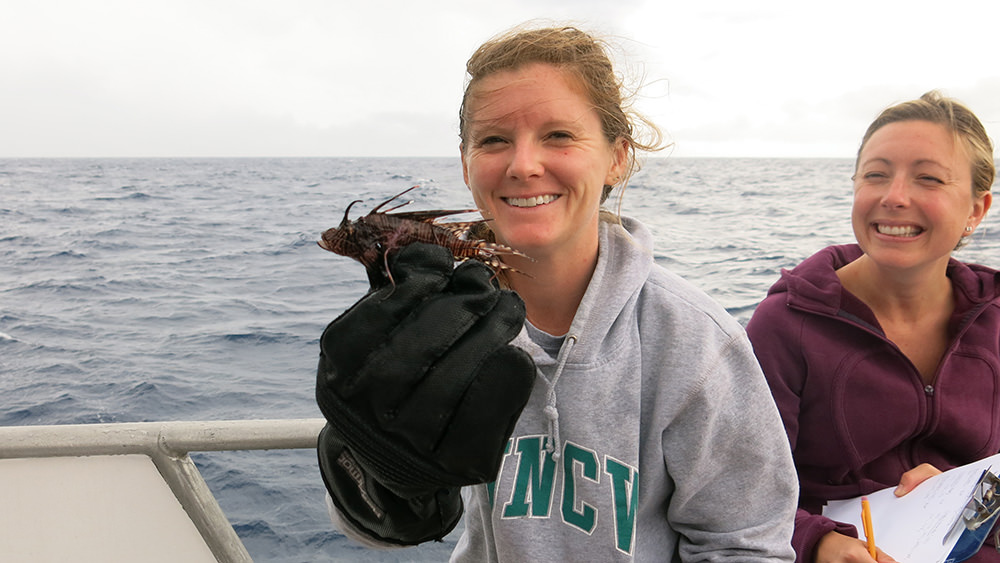 A woman holds up a small lionfish using a puncture proof glove, as another woman looks on.