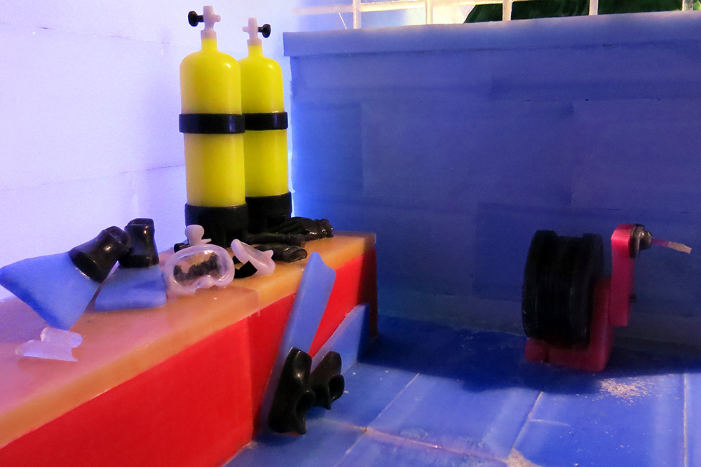 Ice replicas of two sets of scuba gear, including tanks, masks, fins, and gloves, arranged on a bench of the ice R/V MANTA. An ice replica of a small winch is also visible on the deck.
