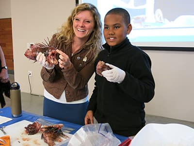 Michelle Johnston holding up a headless lionfish carcass while a student helper holds up the lionfish head
