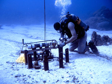 A diver kneeling in the sand next to a railway wheel on the sea floor.  Attached to the railway wheel are water quality instruments.
