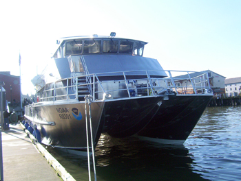 A view of the R/V Manta from the front as she sits next to a wooden dock.  Lines run from her bow to the dock.  A NOAA logo is visible on the front left of the boat and her shiny aluminum sides are reflecting the glare of the sun. 