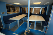 The main dining area of the vessel with two tables on pedastal legs with bench seating surrounding them.  Windows to the outside are visible behind the tables, with another window to the left that looks into another interior work space.