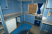 One end of the lab area showing an L-shaped counter with blue cabinets and a small refrigerator beneath them.  A small sink for working with acids is located in the countertop to the right of the fridge and just above a hazardous materials storage unit.  A window above the counter looks outside.ext to blue cabinets.  A small sink for working with acids