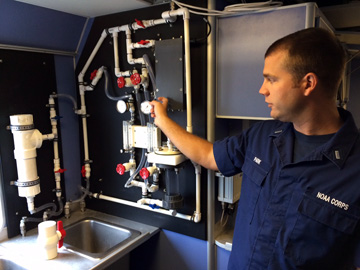 Man pointing out features of a system of water pipes and gauges mounted on the wall above a sink on R/V MANTA