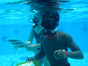 Underwater view of two students in snorkel gear. One is carrying coral ID pictures and the other is carrying a clipboard 