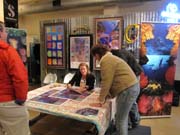 A woman seated at a table autographing posters while other people look on.  Colorful artwork is on display behind her.