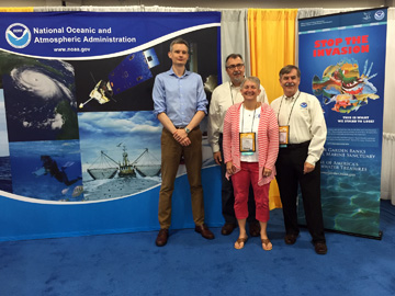 3 men and a woman standing between a NOAA backdrop and a Stop the Invasion lionfish banner in an exhibit booth of a conference hall