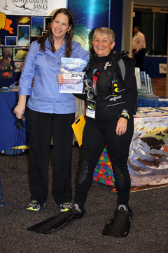 Two women, one holding a book and the other in scuba gear, standing in front of a Flower Garden Banks display at a science teacher conference
