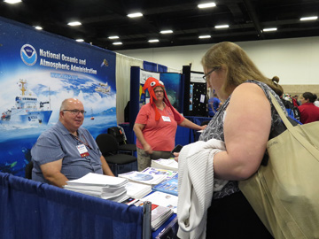 Dennis Cain sitting and Shelley DuPuy standing behind the information tables of the NOAA Education booth while a teacher looks at the selection of materials available.