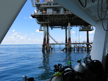 Scuba tanks lined up along the edge of a boat with a gas platform visible just beyond the boat.
