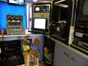 A plush bear toy sits on a stool with a plush sea lion toy in front of all the controls for a remotely operated vehicle.  The controls occupy 3 large cases sitting on a countertop.