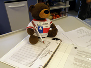 A plush bear toy sits at an open notebook with a pen leaning agains his arm.  The bear is wearing a TX flag t-shirt and a sanctuary volunteer name tag.
