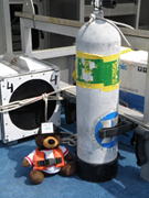 A plush bear toy wearing a hard hat and life jacket sits on the deck of a boat next to a scuba tank.