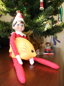 Elf doll holding a foam fish in his lap under a Christmas tree.