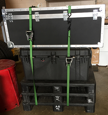 Two black cases strapped on top of three black pallets with a pair of green ratchet straps.