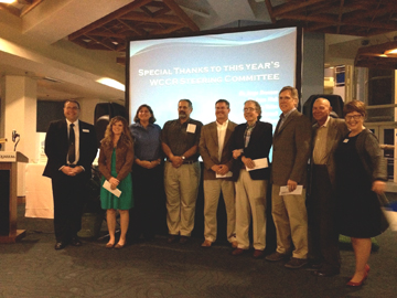 A group of award winners standing in a row in front of a projection screen.