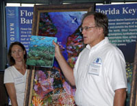 G.P. Schmahl holding up a coffee table book about the Flower Garden Banks while standing in front of a colorful painting of the Flower Garden Banks.  Emma Hickerson is standing to the left.