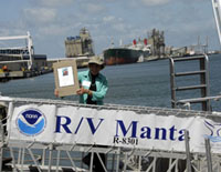 Sylvia Earle standing on the gangway to the Manta.  The NOAA Logo and vessel name are visible ona banner hanging on the side of the gangway.  Sylvia is holding the dedication plaque.