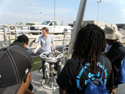 Students listening to a person explaining a large piece of equipment on the deck of a boat.