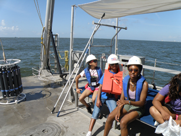 Students sitting on a bench on the deck of a boat near a CTD carousel.