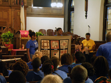 Two students holding up a quilt in front of a church audience.