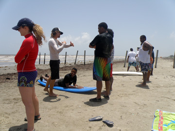 Student lying on surf board on beach while instructor explains surfing technique. Other students stand nearby watching.