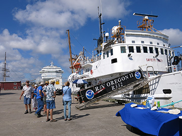 A table with a blue tablecloth sits on the dock next to NOAA ship Oregon II. Guests stand to the left of the ship's gangway waiting to board