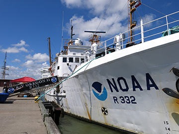 Looking down the starboard side of NOAA ship Oregon II from bow towards stern as it is docked at Pier 21 in Galveston. A gangway leads from the ship to the pier for pedestrian access. NOAA R232 is visible next to the NOAA logo at the bow of the ship.