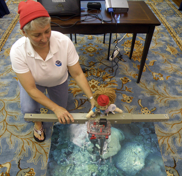 A woman and a doll, both wearing red hats, doing a reef monitoring demonstration with a t-frame