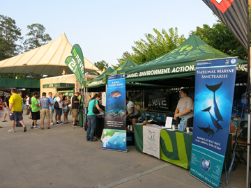 The sanctuary's display booth under a green pop-up shelter that says Music. Environment. Action. Two sanctuary banners are displayed on either side of the shelter.