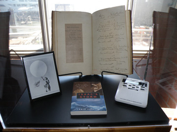A hand-written weather journal from the 1800s in a display case with a photo, a book, and a weather radio.