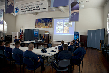 Coast Guard staff seated around tables watching a Powerpoint presentation in a large room