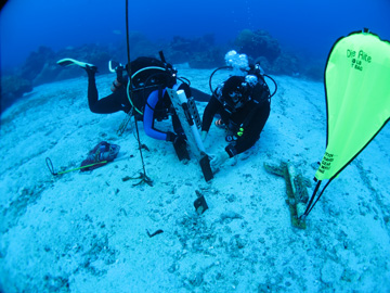 Divers changing out water quality instruments at an underwater station. An inflated lift bag rests on the sand nearby.