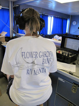 A woman wearing headphones while sitting on a stool with her back to the camera. The back of her shirt says Flower Garden Banks R/V Manta.