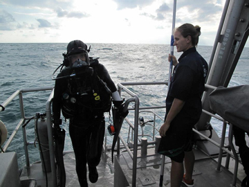 Diver with a full face mask stepping back onto the boat as another person looks on.