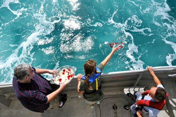 Three people leaning over a boat rail tossing rose petals into the water.