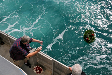 Three people along a boat rail tossing a wreath into the sea.