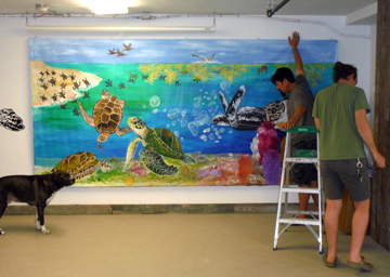 Two men hanging a large sea turtle mural on a wall as a dog watches.