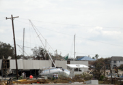 A large sailboat and oher boats on land next to a damaged building.  People standing next to the sailboat are smaller than the keel.