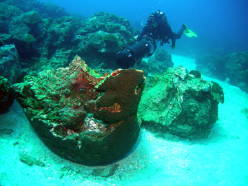 A large boulder of brain coral lies upside down in the sand.  A diver with an underwater scooter hovers just above and to the right of the boulder.