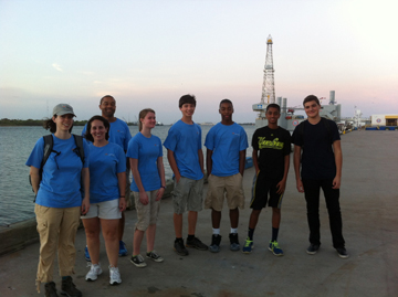 Group of people standing on a pier in Galveston with an oil rig derrick visible behind them.