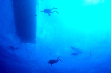 Looking up at the underside of a boat moored at the surface.  Silhouettes of 4 scuba divers are visible near the boat.