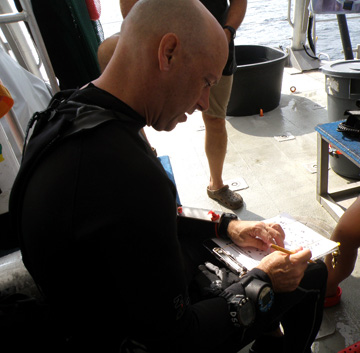 Kevin dressed in his wetsuit, sitting on a dive bench, reviewing the dive plan on his clipboard