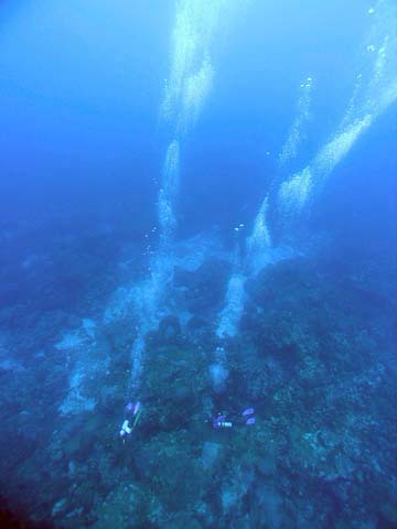 Underwater and looking down on two scuba divers swimming over a coral reef far below.  Streams of bubbles are rising from each of the divers.