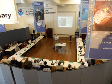 Looking down on a meeting from an overhead balcony.  Meeting participants are seated around tables placed in a large U shape.