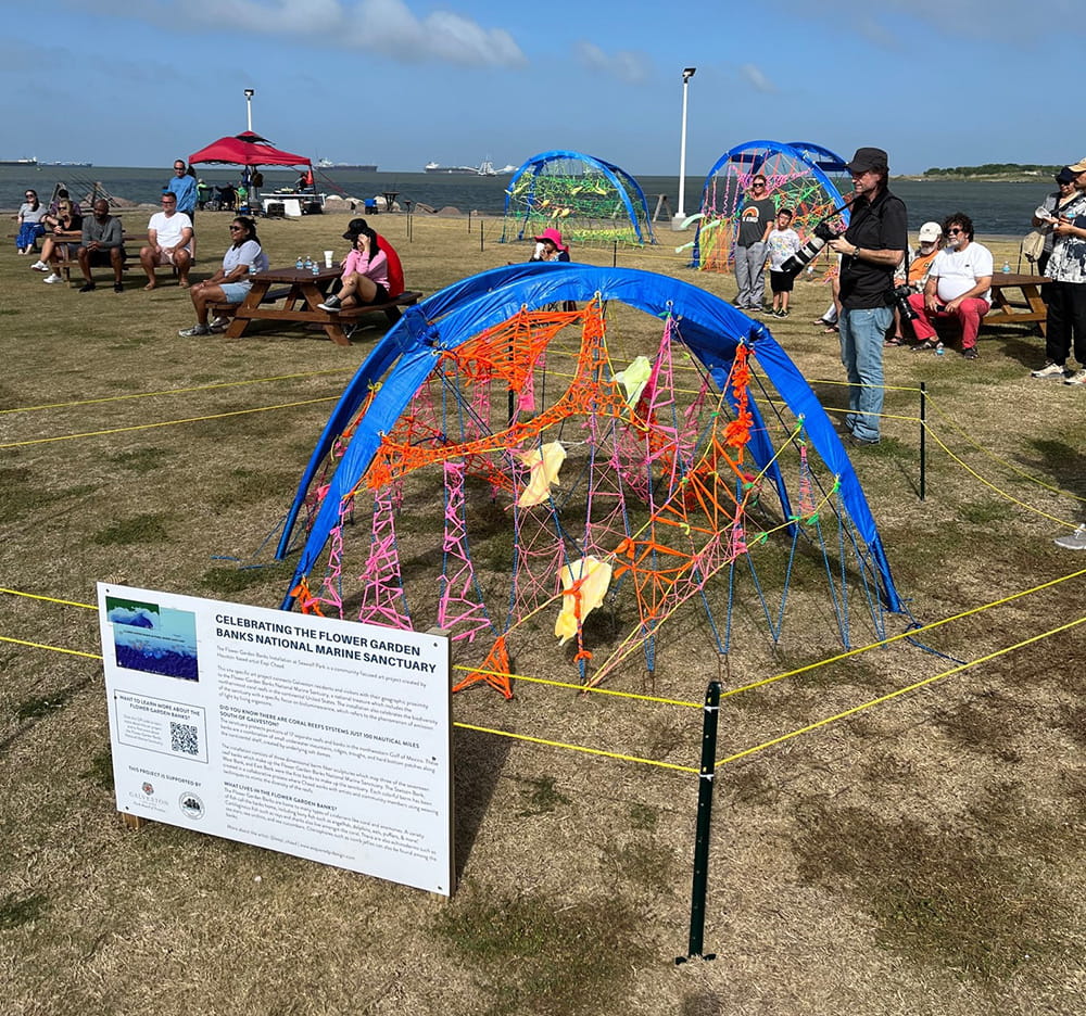 An outdoor art project that looks like 3 camping tent frames covered in colorful ribbons and string spaced out across a grassy area between picnic tables.